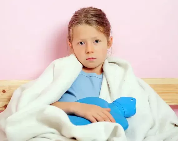 Ein Mädchen sitzt in eine Decke gehüllt und mit Wärmflasche auf dem Bauch im Bett. 